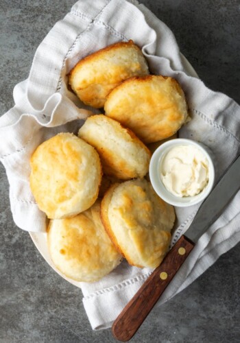 Top view of baked 7UP biscuits in a cloth-lined dish, with a small bowl of butter and a butter knife.