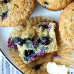 Overhead view of one half of a banana blueberry muffin resting in a muffin liner, next to more muffins on a plate.