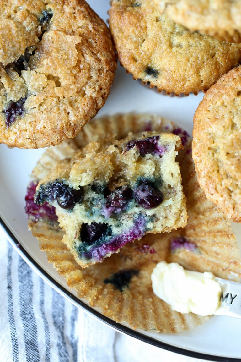 Overhead view of one half of a banana blueberry muffin resting in a muffin liner, next to more muffins on a plate.