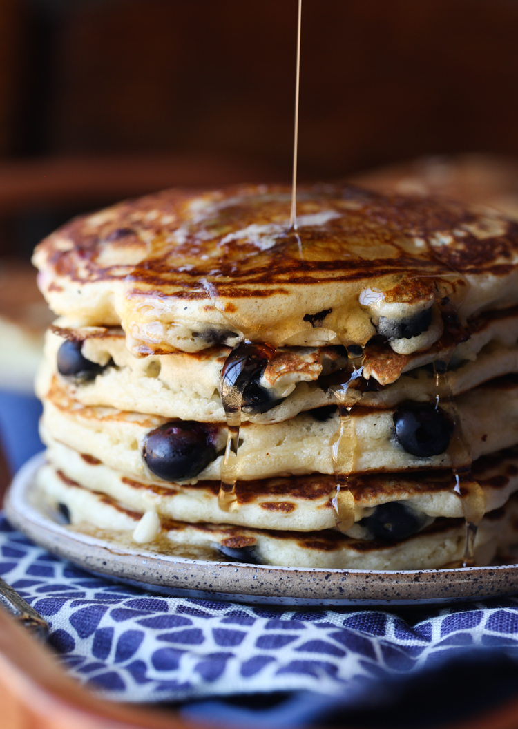 A stack of blueberry pancakes on a plate, being drizzled with maple syrup.