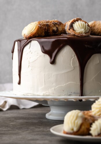 Cannoli cake on a cake stand drizzled with chocolate ganache and garnished with cannoli pastries.