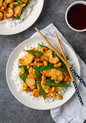 Overhead view of cashew chicken served over a bed of jasmine rice on a plate, next to chop sticks and a bowl of soy sauce.