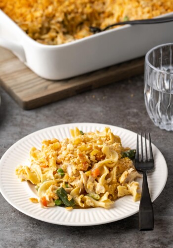 A serving of chicken noodle casserole on a white plate with a fork, with the casserole in the background.