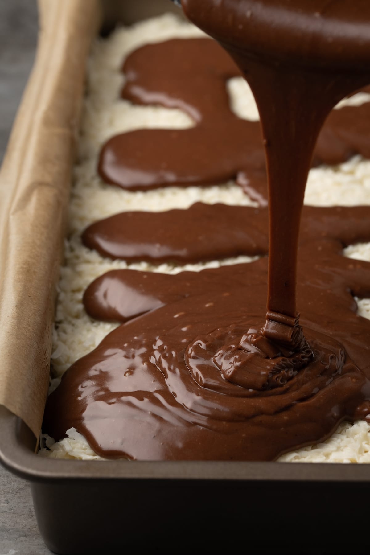 Chocolate icing is poured over top of the brownie and coconut layers in a parchment-lined baking pan.