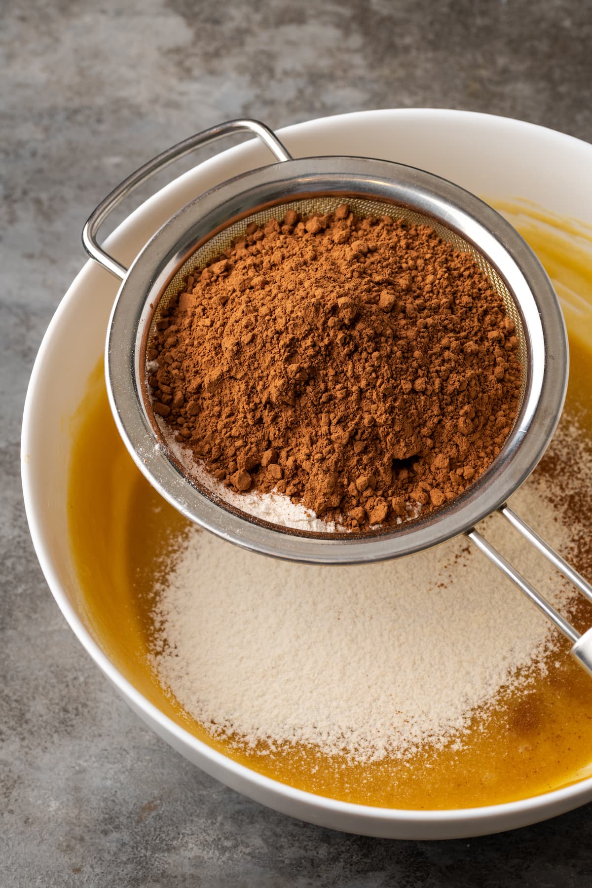 Dry ingredients sifted through a fine mesh sieve over a bowl of brownie batter ingredients.