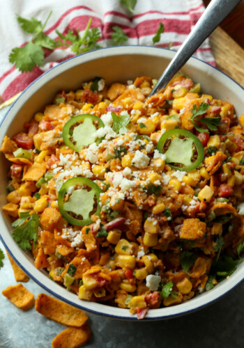 Spicy Frito corn salad topped with cotija cheese and sliced jalapeños in a large white salad bowl with a serving spoon.