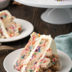 A slice of frosted confetti cake on a white plate, with a second slice on a plate next to a cake stand in the background.