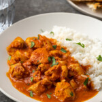 Instant pot butter chicken served on a white plate next to a side of basmati rice, garnished with fresh chopped cilantro, with a plate of naan bread in the background.