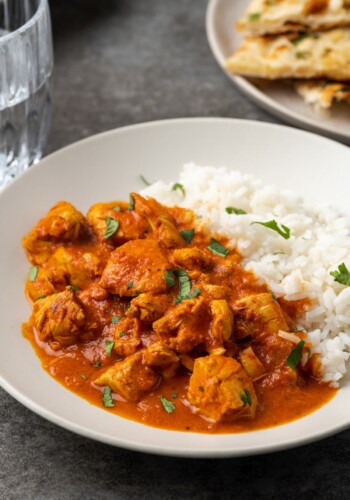 Instant pot butter chicken served on a white plate next to a side of basmati rice, garnished with fresh chopped cilantro, with a plate of naan bread in the background.