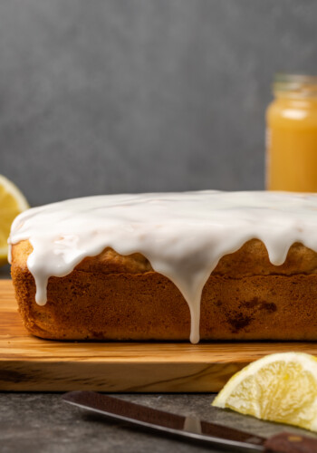 Lemon pound cake topped with lemon glaze on a cutting board, next to a jar of lemon curd and lemon wedges.