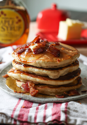 A stack of Maple Bacon Pancakes topped with butter, more chopped bacon, and maple syrup