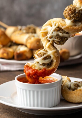 A meatball sub on a stick is dipped into a ramekin of marinara sauce.