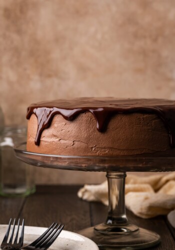 A frosted chocolate layer cake topped with chocolate ganache on a cake stand, next to a white plate with two forks.