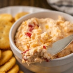 Close up of a bowl of pimento cheese served with a cheese knife on a platter of crackers.