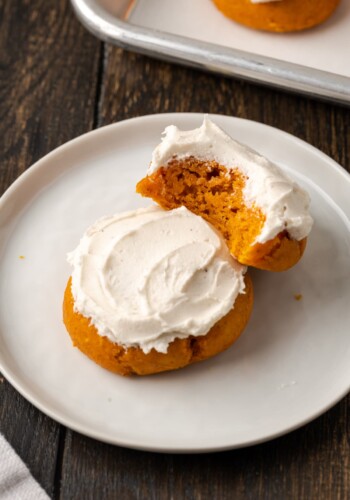 Two frosted cake mix pumpkin cookies on a white plate, with a bite missing from one cookie.