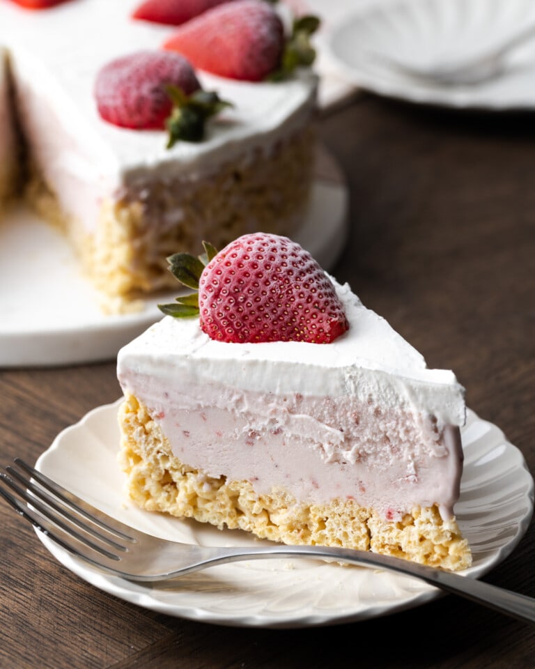 A slice of Krispie Treat ice cream pie on a white plate next to a fork, garnished with a fresh strawberry, with the rest of the pie in the background.