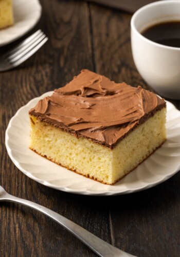 A slice of frosted sour cream coffee cake on a white plate next to a fork and a cup of coffee.