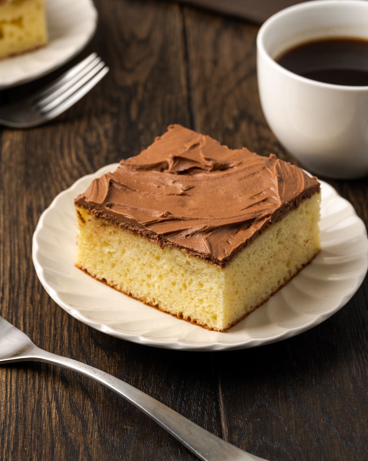 A slice of frosted sour cream cake on a white plate next to a fork and a cup of coffee.