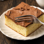 A fork cuts into the corner of a slice of frosted sour cream coffee cake on a white plate.