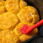 A rubber spatula brushing melted butter over baked sweet potato biscuits in a cast iron skillet.