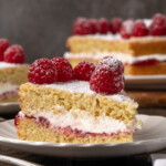 A slice of Victoria sponge cake on a white plate, topped with fresh raspberries, with the rest of the cake on a plate in the background.